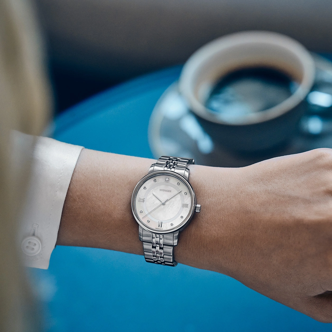 A woman wearing a silver Wenger watch with coffee nearby.