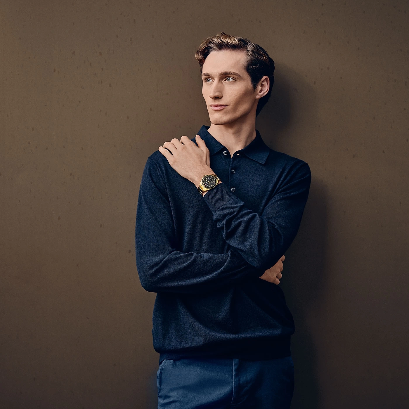 Man wearing a watch, leaning against a brown wall, looking thoughtful and stylish.