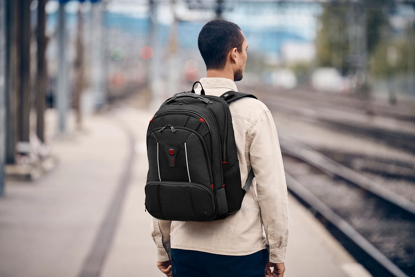 Man with backpack on train station platform, looking at tracks.